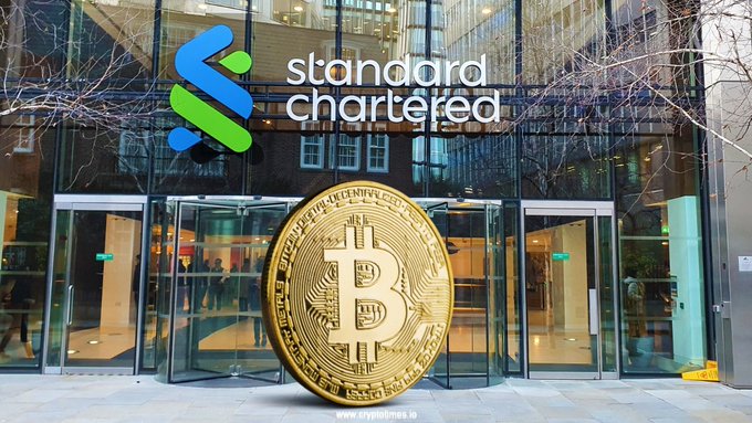 A large golden Bitcoin sculpture stands prominently in front of a Standard Chartered bank branch entrance, symbolizing the bank's recent launch of BTC and ETH trading services for eligible UAE investors through their existing bank accounts. The image visually reinforces the post's announcement of Standard Chartered integrating cryptocurrency custody and trading directly into traditional banking.