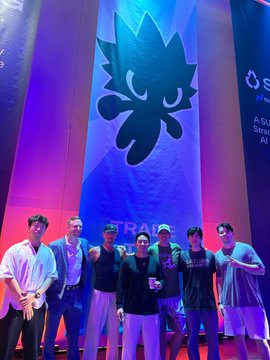 A group of six people pose together on stage in front of a large blue banner displaying a spiky-haired mascot logo and the text 'A Sum... Stra... A...'.