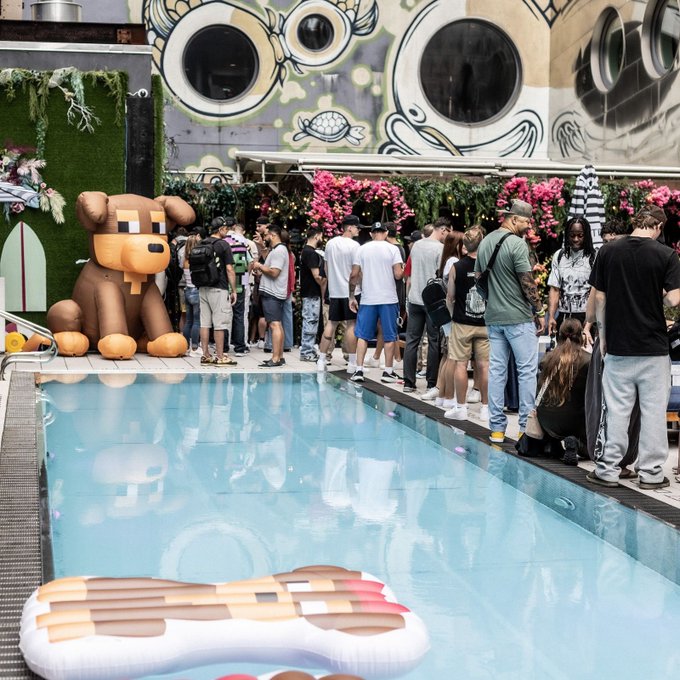 An outdoor pool party scene with a large inflatable dog, an oversized dog bed, and a crowd of people gathered near a wall with graphic mural art.