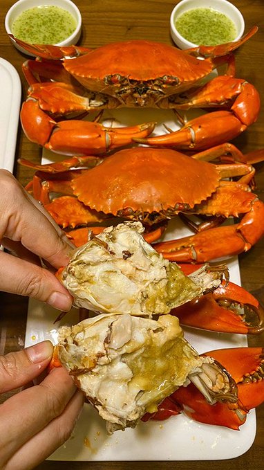 A close-up of a meal featuring two whole cooked crabs with bright orange-red shells on a white plate, with one crab cracked open to reveal its meat and roe, accompanied by two small bowls of green dipping sauce.