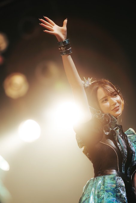 A performer wearing a teal and black stage costume with a small crown waves at the camera, backlit by bright stage lights against a dark background.