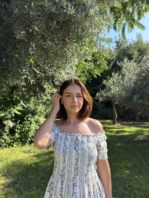 A young woman with shoulder-length brown hair wearing a white off-shoulder floral dress, posing outdoors in a sunlit garden with olive trees in the background.