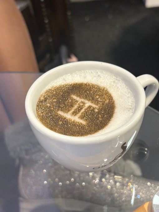 A white coffee cup on a glass table contains a beverage with a triangular symbol etched into the foam.