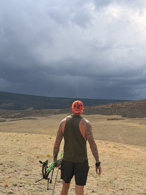 A man wearing an orange "Georgia" cap and olive tank top stands on a grassy hillside holding a compound bow, gazing out at a vast landscape beneath dark, stormy clouds. The scene captures a focused "locking in" moment as he prepares to hunt under dramatic skies.
