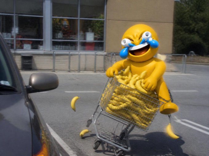 An advertising mascot in a yellow costume with a crying-laughing emoji face pushes a shopping cart full of bananas in a parking lot.