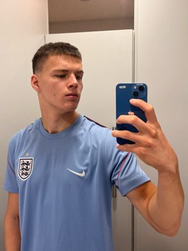 A young man takes a mirror selfie wearing a light blue England national football team jersey with the Three Lions crest and Nike logo.