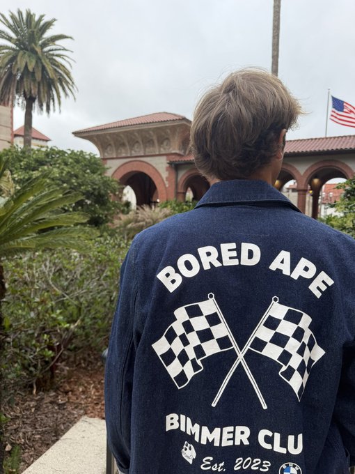 A person photographed from behind wearing a dark blue jacket with a large white graphic of crossed racing flags and text reading 'BORED APE BIMMER CLUB Est. 2023', standing outdoors with a palm tree and American flag visible.