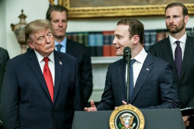 President Donald Trump stands beside another man speaking at a podium bearing the Presidential Seal in what appears to be the White House.