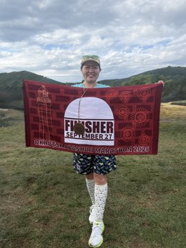 A runner proudly holds up a "FINISHER SEPTEMBER 27" banner from the Hoka "Mt Disappointment Endurance Run 2023," celebrating their achievement while standing in a grassy field with mountains in the background—likely sharing their fitness accomplishment with the #STEPN community.