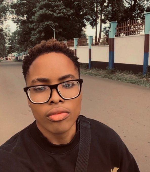 A selfie of a young person wearing black-rimmed glasses and a black shirt, standing on a street with a fence and trees in the background.