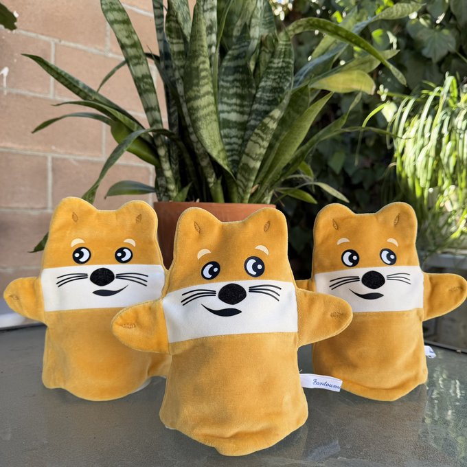 Three yellow fox-shaped plush toys sit on a counter in front of a snake plant and other greenery.