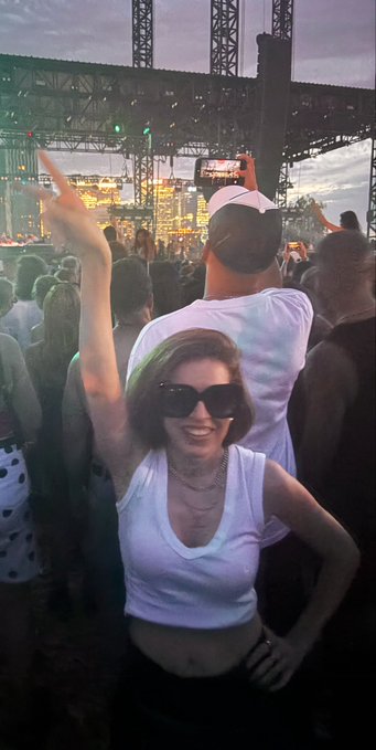 A woman wearing sunglasses and a white tank top smiles while dancing at an outdoor concert festival.