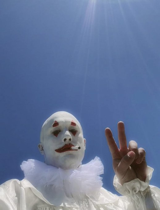 A clown wearing white face paint with red diamond markings above the eyes, a red painted smile, a ruffled collar, and a cigarette in mouth makes a peace sign against a bright blue sky.