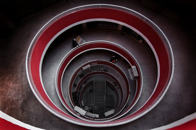 A top-down view into a striking spiral atrium with red and white railings, showing multiple levels descending toward the bottom.