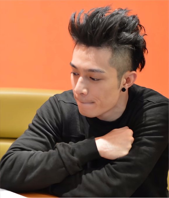 A young man with spiky black hair and an undercut hairstyle poses in a black long-sleeve shirt against an orange wall, wearing a black earring.