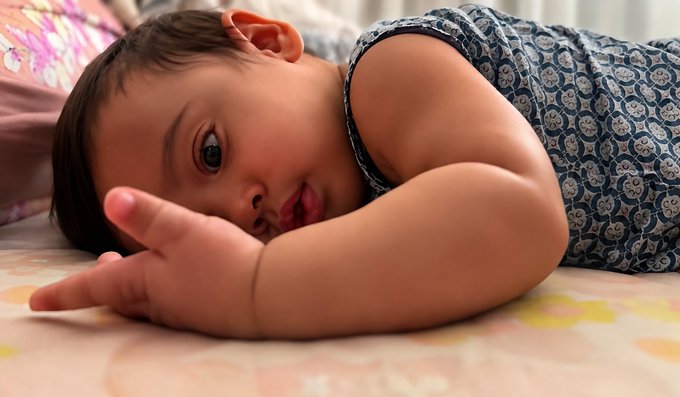 A close-up of a baby wearing a blue patterned sleeveless top, lying on a bed with pink floral sheets and looking at the camera.