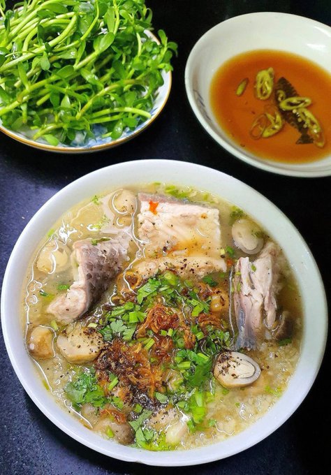 A bowl of Vietnamese-style noodle soup, likely pho, garnished with fried shallots, cilantro, and black pepper, served with a side of fresh herbs/watercress and a small dish of dipping sauce with chili.