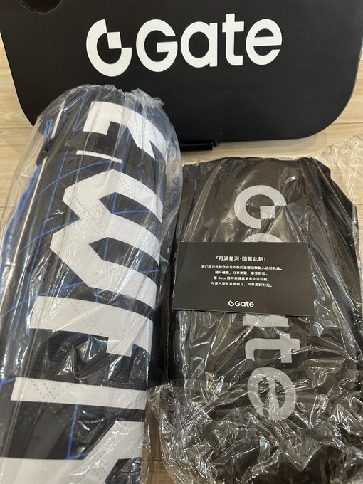 Two rolled black items (possibly mats or banners) wrapped in plastic bearing the "Gate" logo, with a black bag and an informational card also displaying the Gate logo in the background.