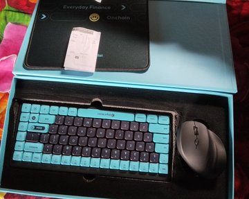 An open teal and black keyboard and mouse set rests inside its box packaging, with a tablet displaying text visible in the background.
