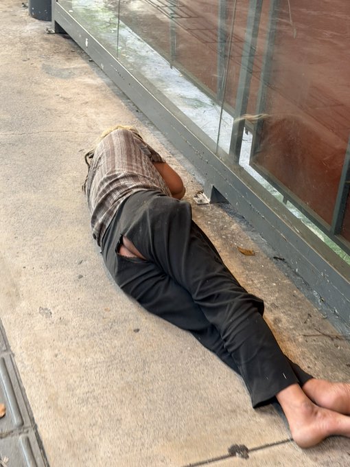 A person is lying face down on a concrete sidewalk next to a glass-walled structure, wearing a striped shirt and black pants with a rip in the back pocket, and is barefoot.