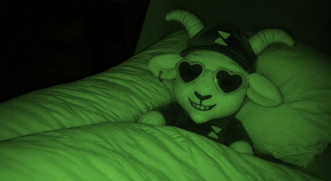 A stuffed goat toy wearing heart-shaped sunglasses and a cap, lying in bed under a green night vision filter.