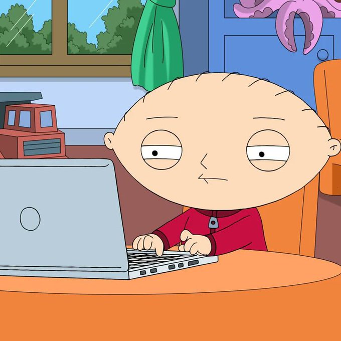 Cartoon character Stewie Griffin from Family Guy typing on a silver laptop while sitting at a table in a room.