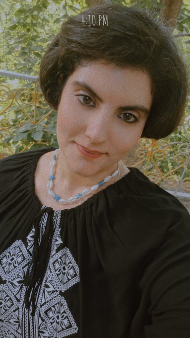 A selfie of a woman with short dark hair wearing a black embroidered top and a blue-beaded necklace, with "4:10 PM" displayed on the screen.
