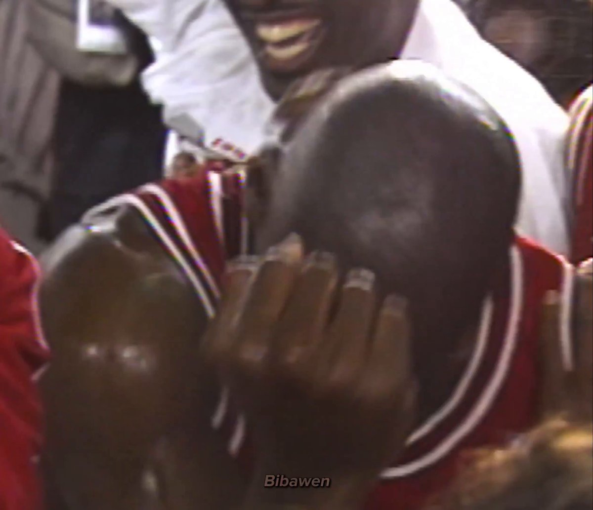 The image shows a man wearing a red basketball jersey, with his head tilted back as he looks down or laughs. He has short hair and is smiling for the camera. The background of the photo includes other people present but they are blurred out, making them less noticeable compared to the main subject in focus.