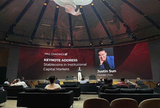 A speaker delivers a keynote address at a Hall Chadwick event, with a large screen displaying "Stablecoins in Institutional Capital Markets" by Justin Sun, Founder of Tron. The image connects to the post by highlighting Tron founder Justin Sun speaking at a financial industry conference, contextualizing the launch of TRON's EnergyBox platform as part of the broader ecosystem's expansion into institutional finance.