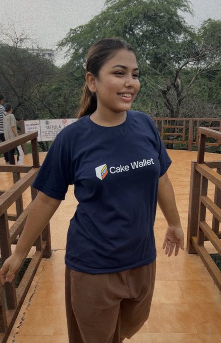 A young woman wearing a navy blue t-shirt with the "Cake Wallet" logo smiles while standing outdoors on a wooden walkway surrounded by trees.