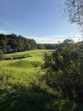 A scenic view of a green golf course fairway bordered by trees, with a sand bunker visible and a partly cloudy blue sky above.