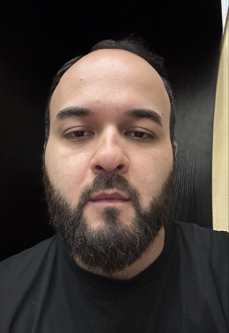 A close-up portrait of a man with dark hair and a beard, looking directly at the camera with a neutral expression against a dark background.