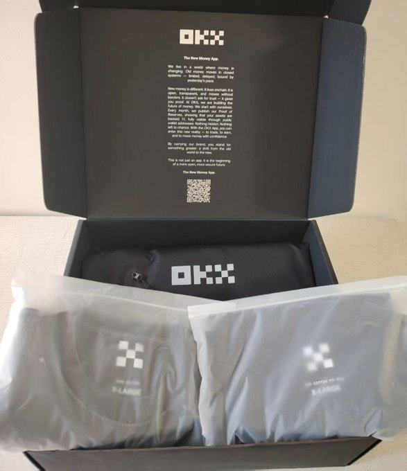 An open black OKX "The New Money App" promotional box reveals neatly folded dark clothing items in plastic packaging.
