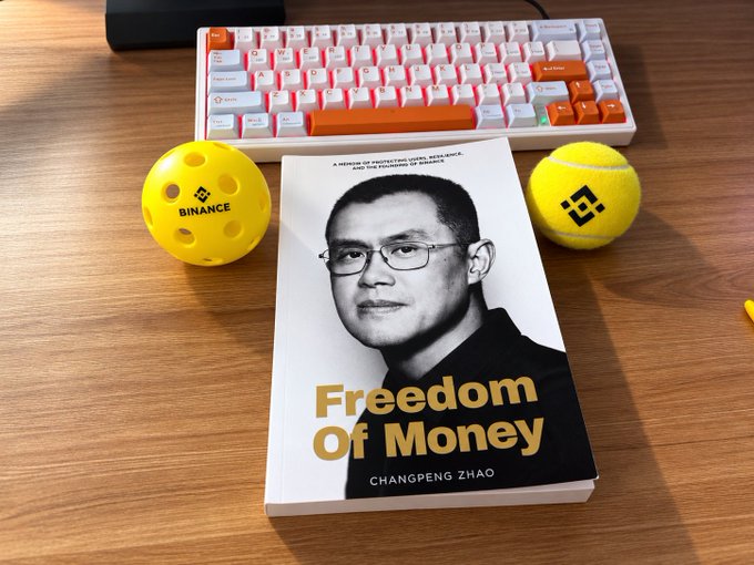 A book titled "Freedom of Money" by Changpeng Zhao placed on a wooden desk between two yellow Binance-branded balls and in front of a white mechanical keyboard.