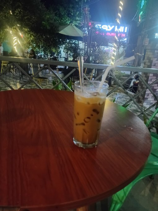 A glass of iced milk coffee with a straw sits on a wooden table at an outdoor nighttime venue, with neon signs visible in the background.