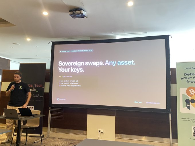 The image shows a man standing behind a large screen, giving a presentation or speech about Bitcoin technology. The screen displays information related to Bitcoin L2s, which are virtual wallets used for storing digital currency. The man appears to be explaining the concept of Sovereign Swaps and how they can be applied to Bitcoin L2s. There is also a laptop on the table in front of him, possibly being used as part of his presentation or demonstration.