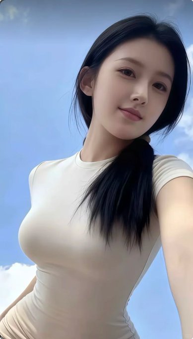 A selfie of a young woman with long black hair wearing a fitted cream-colored t-shirt against a blue sky background, showcasing the stunning beauty referenced in the enthusiastic post praising top-tier looks.