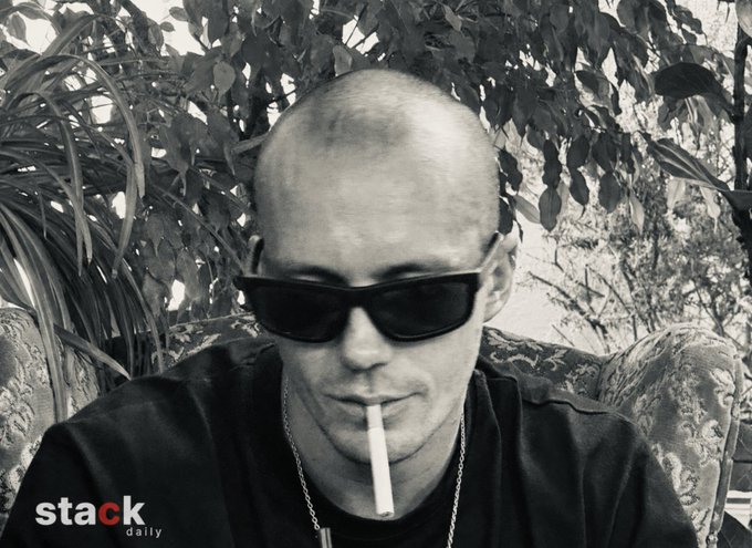 A black and white photograph of a young man with a shaved head wearing dark sunglasses and a t-shirt, smoking a cigarette, with 'stack daily' watermarked in the bottom corner.