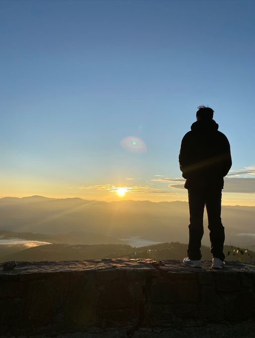 A silhouette of a person standing on a stone ledge overlooking a vast mountain landscape at sunrise, with the sun casting golden rays across the sky and misty valleys below.
