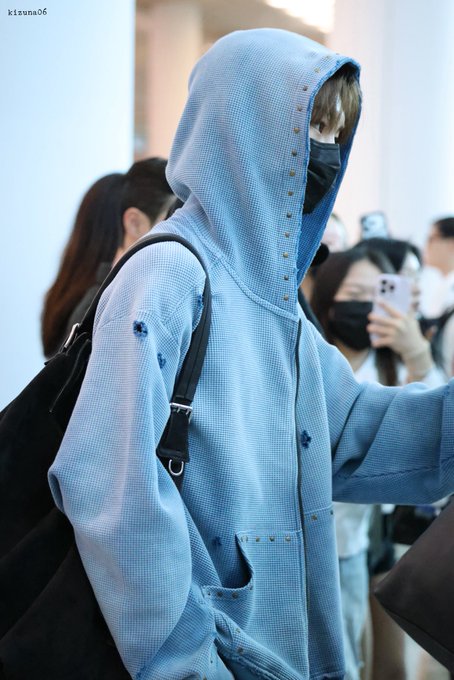 A person wearing a light blue hooded denim jacket with studs and a black face mask, carrying a black bag, walks through a crowd where others are taking photos. The watermark "kizuna06" appears in the top left, and the image aligns with the post about Ahn Shin (안신) arriving at ICN, referencing "ALD1" and "ALPHADRIVEONE."