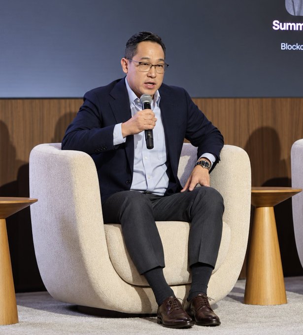 A man wearing glasses, a navy blazer, light blue shirt, and dark trousers speaks into a microphone while seated in a cream-colored armchair at what appears to be a "Summit" panel discussion, with "Blockc..." visible in the background signage, identified as _jikim of the crypto_council commenting on the optimistic progress of the Clarity Act vote.