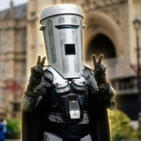 A figure in futuristic metallic armor with a rectangular helmet making peace signs with both hands in front of an ornate building.