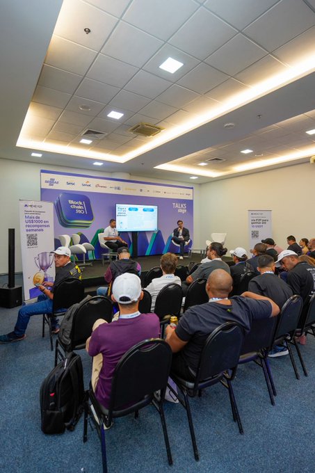 An audience seated in a conference room attends a 'TALKS' session at what appears to be a NEO blockchain event, with two speakers seated on stage.