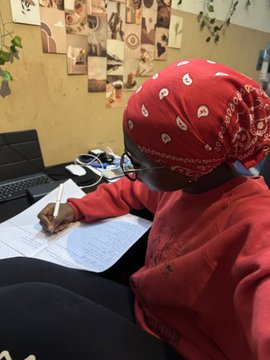 A woman wearing a red bandana and hoodie writes in a notebook at her desk, likely studying for the exams she mentions in the post as the reason for her reduced trading activity this month.