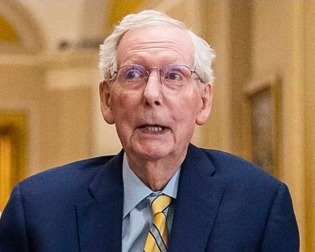 An elderly man with white hair and glasses, wearing a dark blue suit, light blue shirt, and a yellow striped tie, photographed indoors.