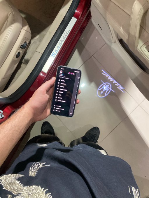 First-person view of someone holding a smartphone displaying a chat or social feed, standing next to a red Maserati car with its logo projected onto the ground.