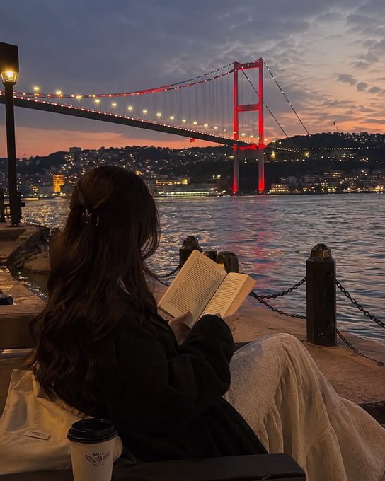 A serene view from behind a woman with long dark hair reading a book by the water, with the illuminated Bosphorus Bridge lit in red against a sunset/twilight sky in the background, possibly in Istanbul.