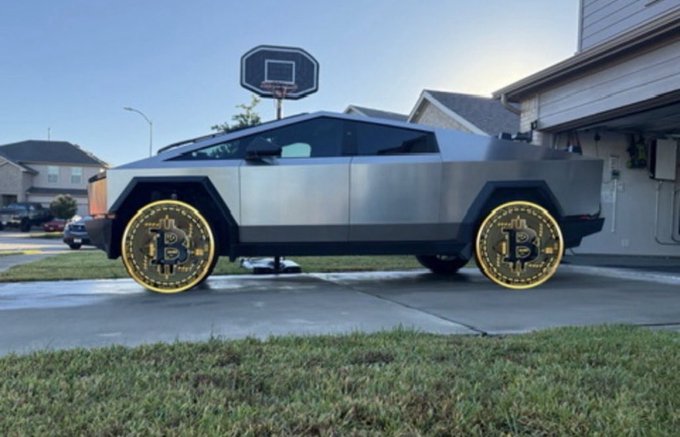 A silver Tesla Cybertruck parked on a residential driveway with its wheels replaced by large gold Bitcoin coin rims.