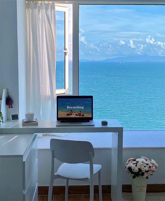 A serene home office setup by a window with an ocean view, featuring a white desk, a laptop displaying 'Recording,' a gray chair, and a vase of flowers on the floor.