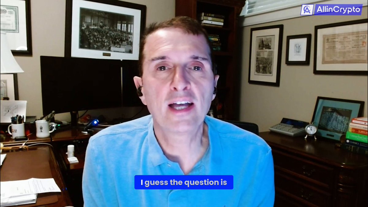 The image shows a man wearing a blue shirt sitting at a desk in front of a computer screen. The background features a wooden desk with various items such as books, a cup, and a keyboard. There are also pictures hanging on the wall behind him. The main focus is on the man's face, which appears to be looking directly into the camera.